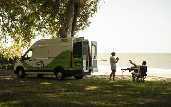 JUCY Condo parked next to beach with two mates sitting at a camping table behind