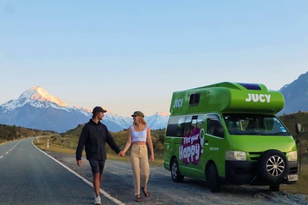 Couple holding hands and walking past a JUCY Condo Campervan parked on the side of the road