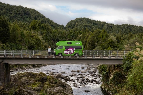 JUCY Chaser campervan parked on NZ bridge overlooking stream with woman driver standing in front