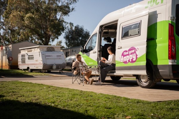 A couple sitting at a camping table beside a parked JUCY coaster campervan hire