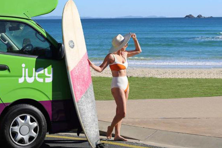 top 5 reasons to hire from jucy in australia 1 Girl with a surfboard next to a JUCY campervan at a beach