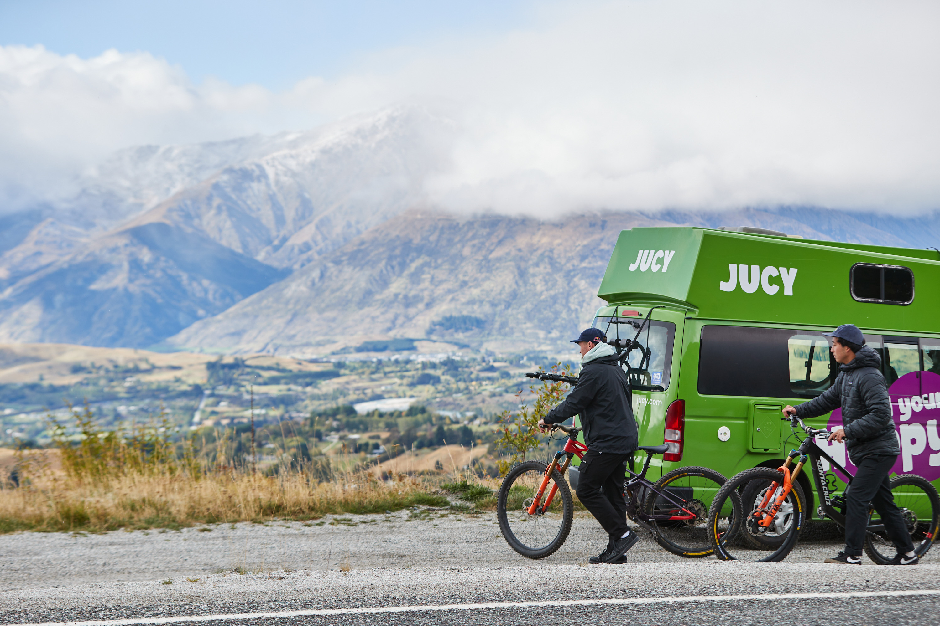 2 friends in winter clothing rolling bikes past a JUCY chaser campervan parked on a hill