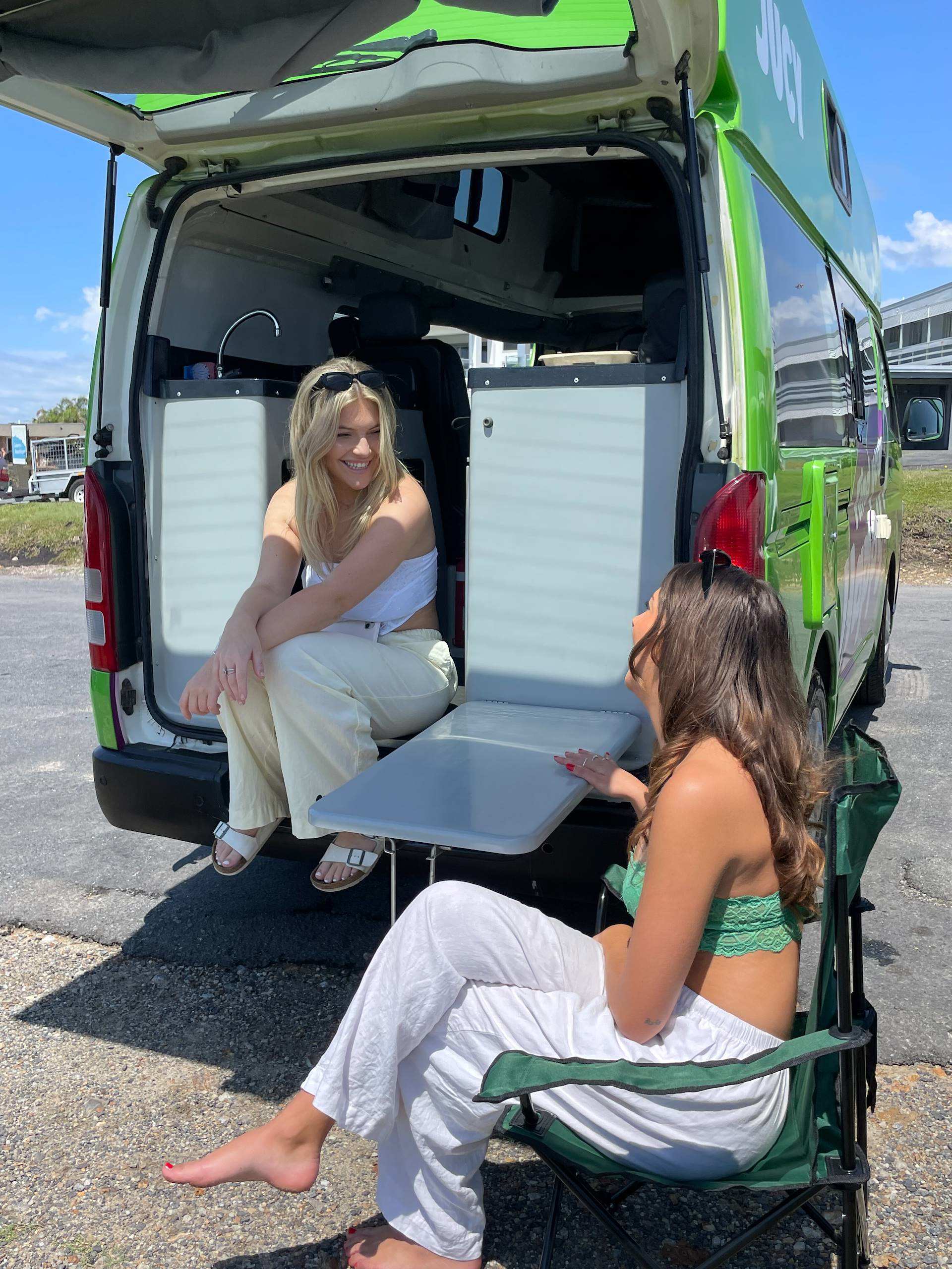 Two girls sitting out the back of a JUCY Condo campervan parked on gravel