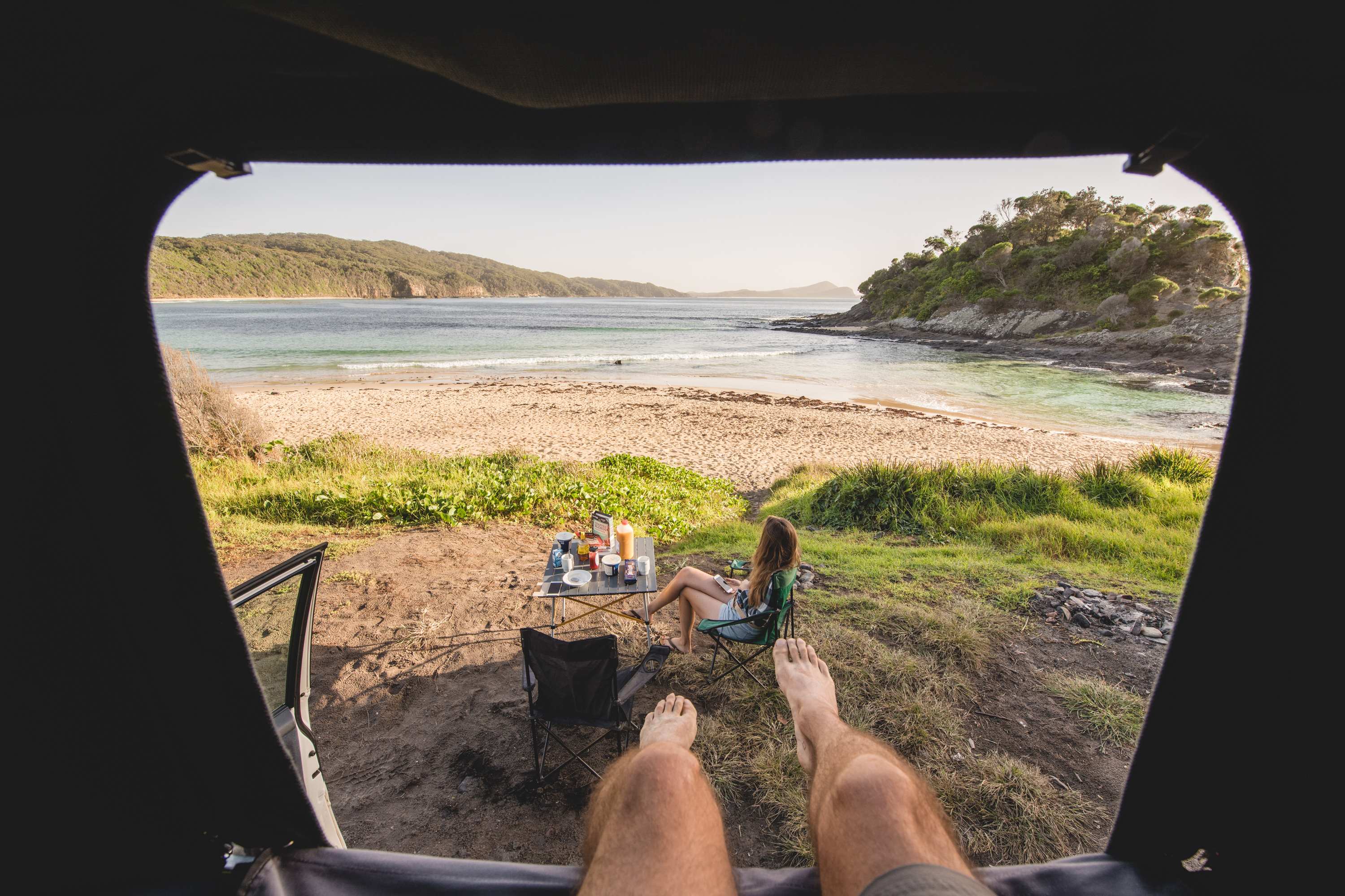 Couple parked next coastal river with camping table and chair set up