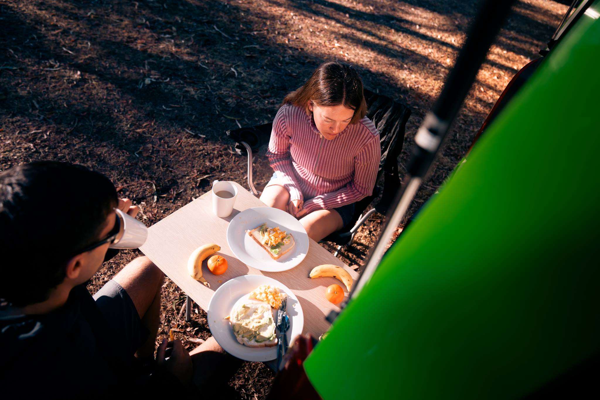 Couple sitting on camping chairs eating breakfast at the back of their JUCY Condo campervan