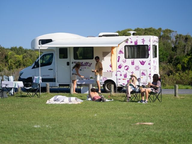 White JUCY Big Kahuna motorhome parked on a green campground with friends sitting under a canopy and socialising outdoors.