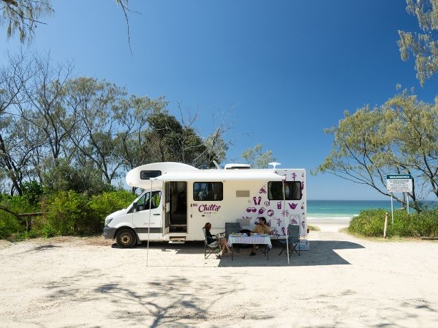 White JUCY Big Kahuna motorhome parked on a beach with white sand and clear blue sky.
